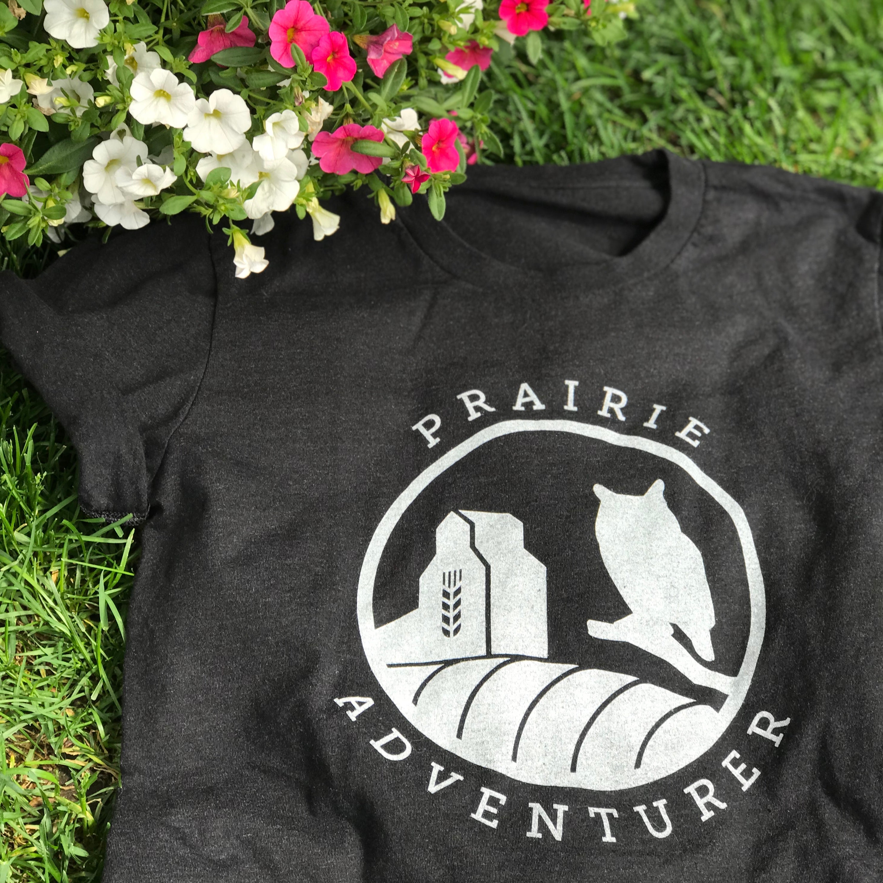 A picture of the Prairie Adventurer tee. This black t-shirt has silhouettes in white of a grain elevator on the left and a Great Horned Owl on the right. The curve of a hay bail appears at the bottom of the graphic.
