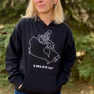 A picture of a woman wearing the Canada Land Mass Hoodie.  The hooded sweatshirt displays the country of Canada outlined in white. The number 9,984,670 appears underneath, as that is the number of square kilometres in Canada.