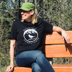A picture of a woman sitting on a park bench wearing the mountain adventurer tee.  This black t-shirt has a white silhouette of a Steller's Jay perched on a branch against a backdrop of mountains with snowy peaks.  
