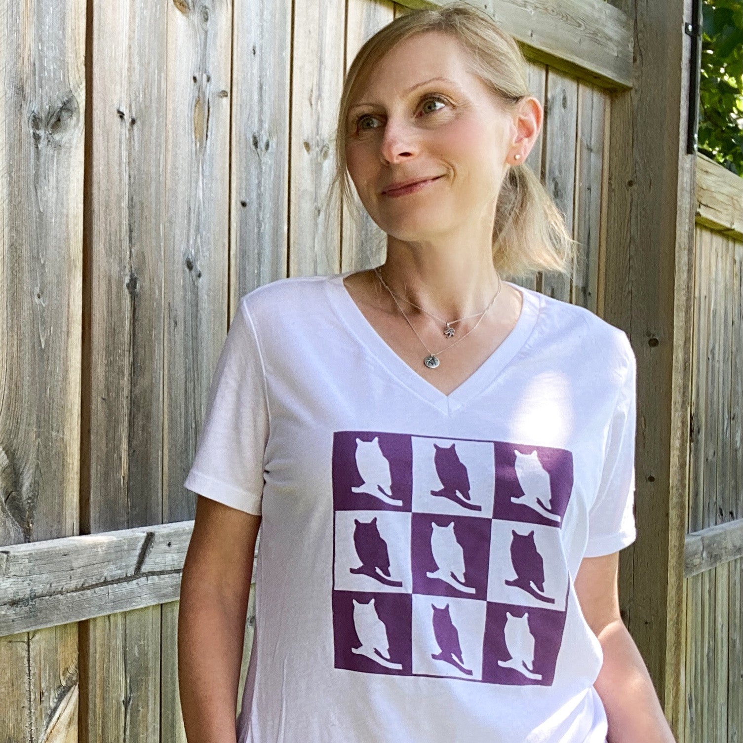 A picture of a woman wearing a white t-shirt.  The image on this t-shirt has nine squares, three per row, of alternating purple and white owls. The white owl has a purple background, and the purple owl is in front of a white background.