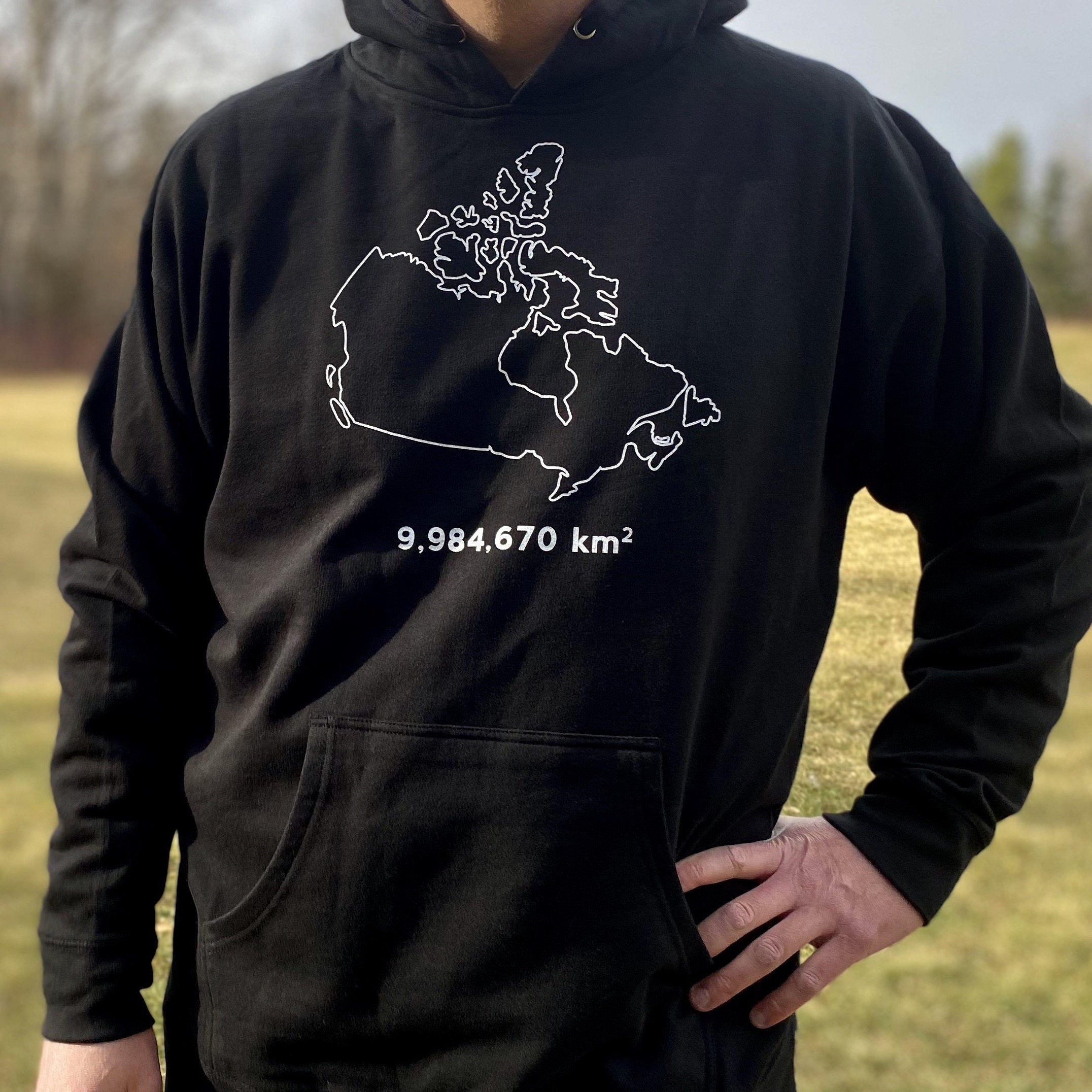 A picture of a man wearing the Canada Land Mass Hoodie. The hooded sweatshirt displays the country of Canada outlined in white. The number 9,984,670 appears underneath, as that is the number of square kilometres in Canada.