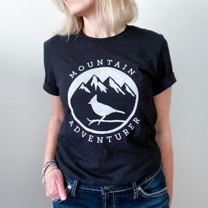 A picture of a woman wearing the mountain adventurer tee. This black t-shirt has a white silhouette of a Steller's Jay perched on a branch against a backdrop of mountains with snowy peaks.