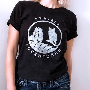 A picture of a woman wearing the Prairie Adventurer tee.  This black t-shirt has silhouettes in white of a grain elevator on the left and a Great Horned Owl on the right.  The curve of a hay bail appears at the bottom of the graphic.